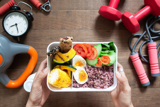 Woman's hands holding lunch box with rice berry, boiled eggs, sweetcorn, pumpkin, tomatoes and cereal bars, Top view with sport and fitness equipments on wooden background