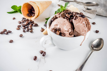 Homemade coffee ice cream, served with coffee beans and mint leaves, with ice cream cones and spoons in the picture. White marble background,