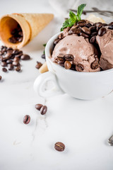 Homemade coffee ice cream, served with coffee beans and mint leaves, with ice cream cones and spoons in the picture. White marble background,