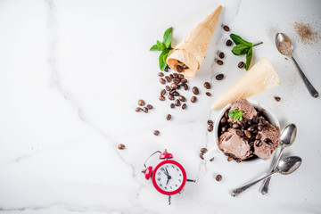 Time for coffee concept, with an alarm clock for hours in the frame. Homemade coffee ice cream, served with coffee beans and mint leaves, with ice cream cones, spoons. White marble background, above
