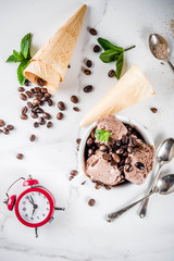 Time for coffee concept, with an alarm clock for hours in the frame. Homemade coffee ice cream, served with coffee beans and mint leaves, with ice cream cones, spoons. White marble background, above