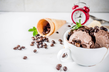 Time for coffee concept, with an alarm clock for hours in the frame. Homemade coffee ice cream, served with coffee beans and mint leaves, with ice cream cones and spoons. White marble background,