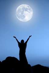 Woman standing on top of big rocks and raising her hands towards the full Moon.