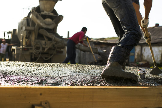 Construction workers are pouring a building foundation. Concrete works with the mixer truck and people with shovels. Mixer truck providing wet cement to the  wooden falsework on the construction site.