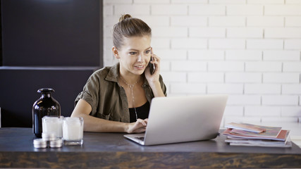 Fototapeta premium A young busy smiling blond casual pretty woman freelancer businesswoman is working at home with a laptop having a call background of a white brick wall.