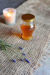 Honey in glass jar, burning candle and lavander flowers on natural linen cloth on grey grunge table background.
