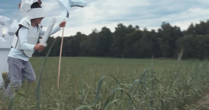 Kid boy wearing cardboard medieval knight armor costume draws a sword from a scabbard near cardboard castle. 4K UHD 60 FPS SLO MO