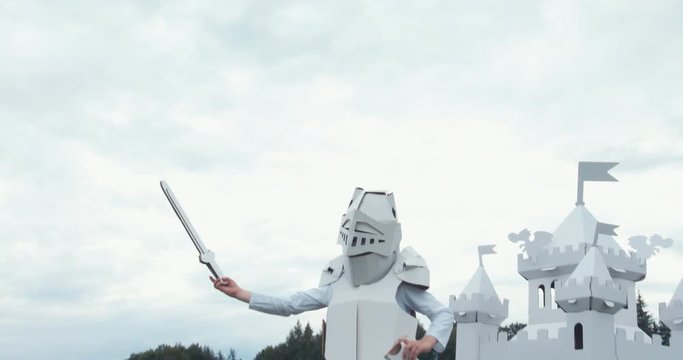 Kid boy wearing cardboard medieval knight armor costume draws a sword from a scabbard against blue sky. 4K UHD 60 FPS SLO MO