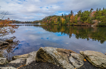 Fototapeta na wymiar Clouds are reflected in a calm Minnewaska Lake in Orange County, NY, surrounded by bright fall foliage on a partly cloudy afternoon