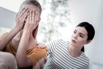 Fototapeta premium Negative emotions. Close up of displeased light haired girl covering her face with hands while her mother keeping hand on her shoulder and supporting her