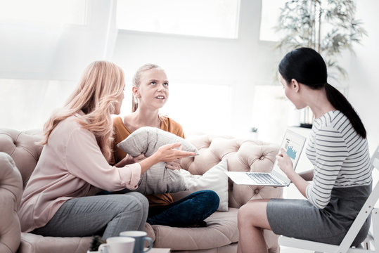 Wrong information. Irritated girl holding pillows and expressing negativity while her mother and psychologist using a laptop and pointing at her problems