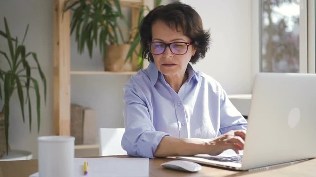 Elderly businesswoman is working with laptop sitting in home office space, successful mature lady wearing glasses is doing job using PC at table in room with daylight. Concept: business person, modern