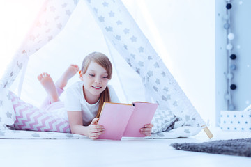 Time for a new adventure. Cute preteen girl smiling while relaxing in a teepee and reading a...