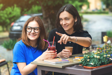 Two young beautiful women friends have a coffee break on terrace, talking with each other pointing aside.