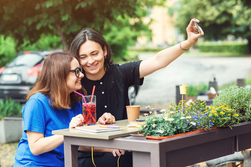 Two smiling girls taking selfie on smartphone, sitting at cafeteria outdoors, have a break together in summer day.