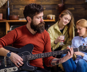 Obraz premium Man with hipster beard playing guitar at college party. Musician rehearsing new song. Bearded man entertaining two blond girls. Guitarist tuning his instrument. Man with calm face composing melody