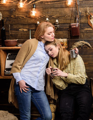 Fototapeta premium Friends spend pleasant evening, interior background. Support and sympathy concept. Friends hugs while have sincere conversation. Girls on relaxed faces hold metallic mugs, enjoy coziness with drinks