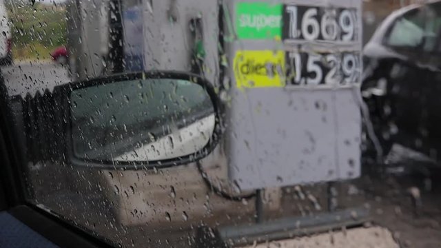POV macchina ni attesa di fare benzina dal benzinaio. Prezzi della benzina sulla destra. Pioggia bagna e schizza i vetri