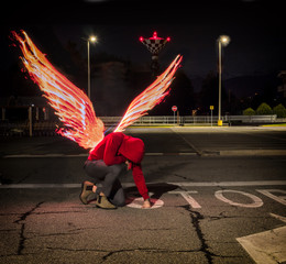 Fallen male angel kneeling in city parking lot, with fire wings spreading from his back....