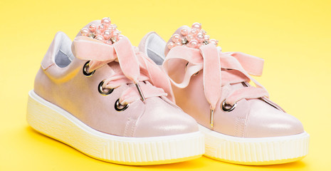 Cute shoes on yellow background. Footwear for girls or women decorated with pearl beads. Pair of pale pink female sneakers with velvet ribbons. Glamorous footwear concept.