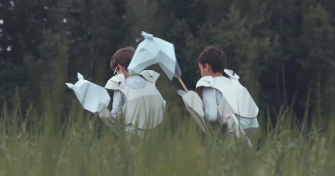 Brothers boys wearing cardboard medieval knight armor costumes riding stick horses through a grass field in summer. 4K UHD 60 FPS SLO MO