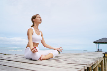 Fototapeta na wymiar Yoga and meditation. Relaxed young woman in lotus pose on wooden deck with sea view.