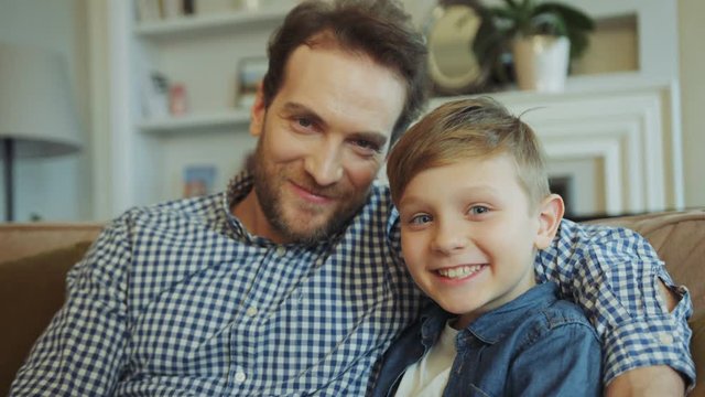 Portrait shot of the smiling happy father with beard abd cute little son sitting on the couch in the living room. Close up. Posing. Indoors