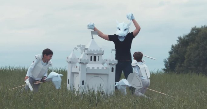Brothers boys wearing cardboard medieval knight armor costumes fighting their father pretending to be a dragon in paper mask. 4K UHD 60 FPS SLO MO