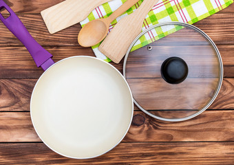 Purple frying pan with glass lid, kitchen wooden utensil and towel on the wooden table. Top view. Cooking meal.