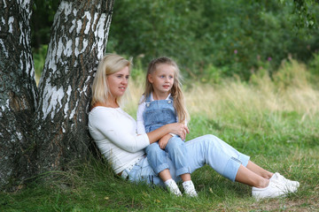 Naklejka na ściany i meble Happy family in nature. Beautiful and happy mother gently hugs her little daughter with blond hair against the background of grass while walking in the summer park. Happy Family Outdoor.