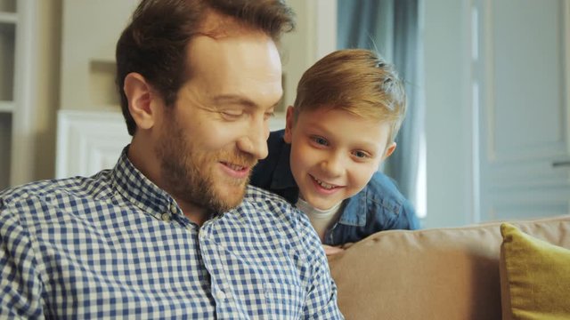 Close up of the cute blonde little boy looking at his father doing something on the laptop computer, both smiling and talking. At home on the couch. Inside