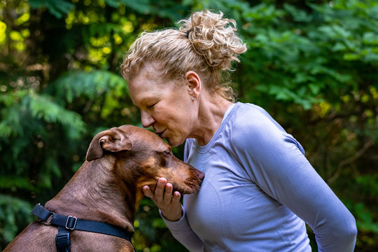 Female blond baby boomer in a purple top in a quiet loving moment with her two tone brown rescue dog out in the woods
