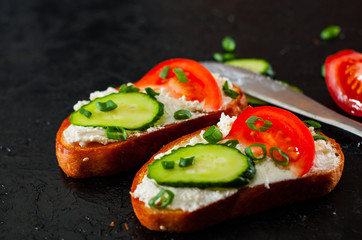 two slice of bread with cream cheese, cucumber, tomato on a black stone board.