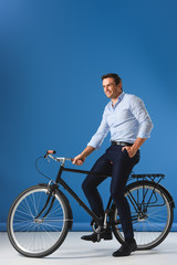 Naklejka premium handsome smiling businessman sitting on bike and looking away on blue