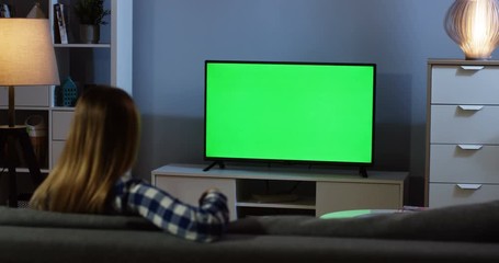 Rear of the blonde woman in the motley blue shirt sitting on the gray couch in the living room, watching TV with green screen and changing channels with a remote control in the evening. Chroma key - Powered by Adobe