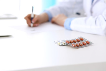 Closeup of pills opposite female doctor filling up prescription form while sitting at the desk in hospital. Healthcare and medicine concept