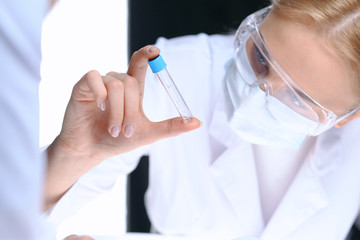 Closeup of scientific research team with clear solution in laboratory. Blonde female chemist holds test tube of glass while her colleague checks results with tablet pc. Blood test, medicine or