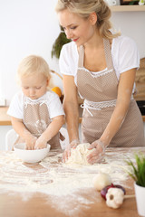 Little girl and her blonde mom in beige aprons  playing and laughing while kneading the dough in the kitchen. Homemade pastry for bread, pizza or bake cookies