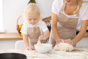 Little girl and her blonde mom in beige aprons  playing and laughing while kneading the dough in the kitchen. Homemade pastry for bread, pizza or bake cookies