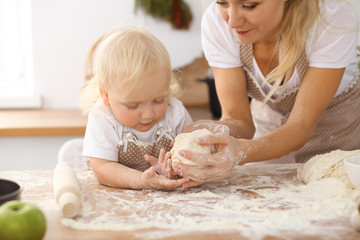 Little girl and her blonde mom in beige aprons  playing and laughing while kneading the dough in the kitchen. Homemade pastry for bread, pizza or bake cookies