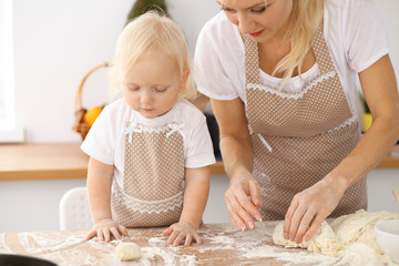 Little girl and her blonde mom in beige aprons  playing and laughing while kneading the dough in the kitchen. Homemade pastry for bread, pizza or bake cookies