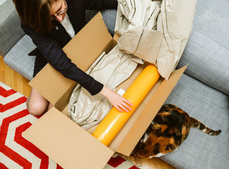 Business woman unpacking unboxing cardboard box box containing yellow yoga zen mat being helped by her pet cat beautiful animal  - obrazy, fototapety, plakaty