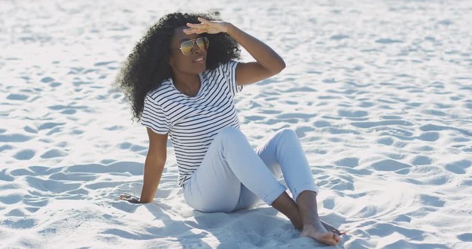 Cheerful African American pretty young woman in sunglasses sitting on the sand and waving helo to somebody. Outside.
