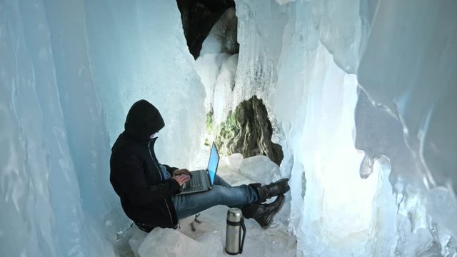 Hacker hacks the server. Man to program on laptop in ice cave. Around the mysterious ice grotto. Man hides himself in a hood. Hacker has criminal cases on the Internet. Guy is warming himself with the
