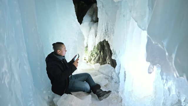 Man is sit on the internet in tablet in an ice cave. Around the mysterious beautiful ice grotto. User communicates in social networks, takes photo and makes selfie on the tablet. He crazy strange