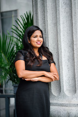 An image of a successful and attractive Indian Asian woman leaning on a concrete pillar during the day. She loks elegant on her black dress and striking a glamorous pose while looking away from the ca