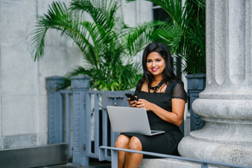 Portrait of a young Indian Asian business woman checking the messages on her phone with her laptop computer on her lap. She is seated in a bench during the day as she works diligently.