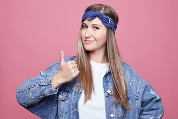 Portrait of attractive self assured young female model keeps thumb raised, shows like gesture, satisfied with something, stands against pink background. People, lifestyle and body language concept.