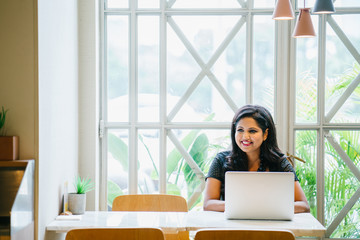 A successful and young desi woman sitting in the cafeteria while working on her laptop. She is wearing a black dress and she is smiling as she looks away from the camera.