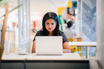 A studio portrait of a youthful but confident and successful Indian woman working on her laptop in a cafeteria.She is sitting down and smiling as she concentrate on her task.